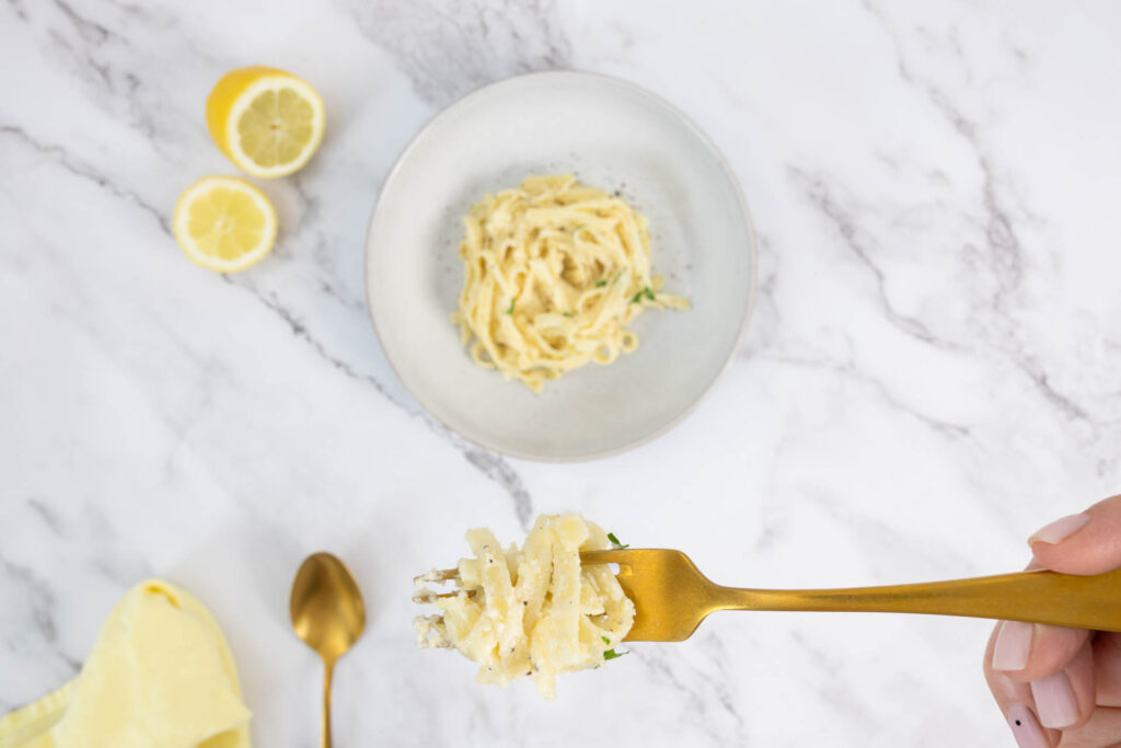 Gabel mit aufgedrehter Zitronenpasta – vegane Tagliatelle al Limone mit frischer Zitrone im Hintergrund