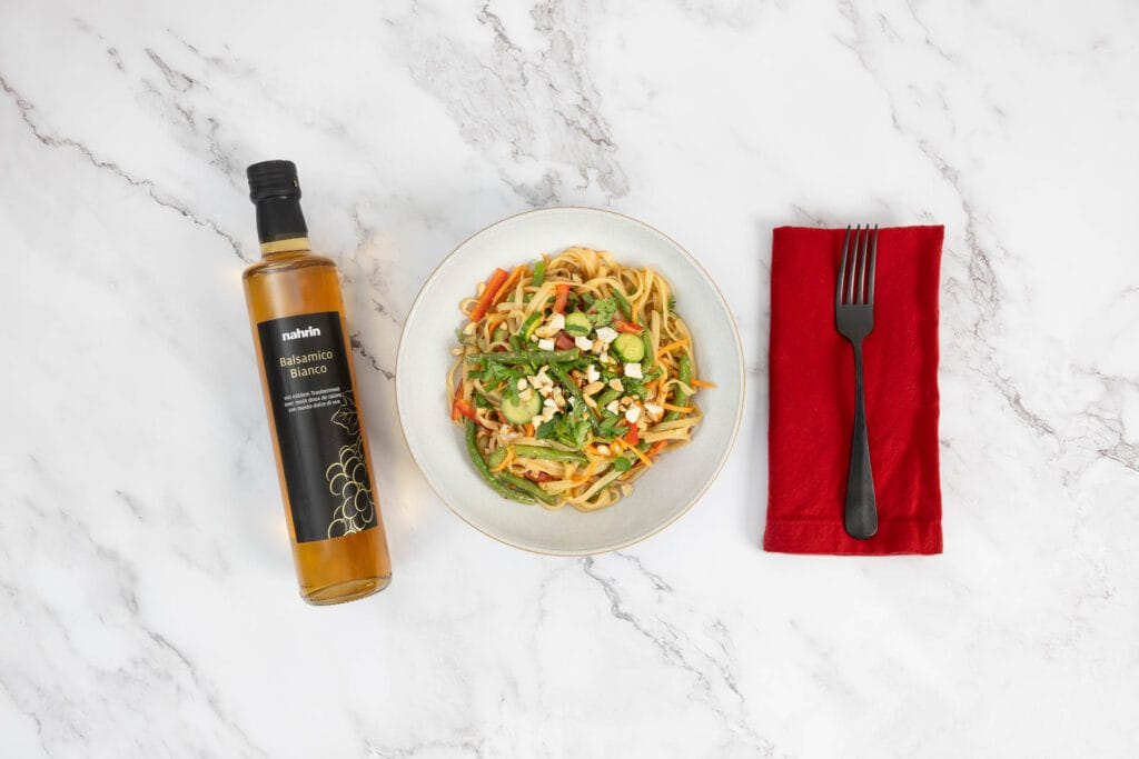 Angerichteter asiatischer Nudelsalat mit grünen Bohnen, Cashews und Dressing, daneben Balsamico Bianco Essigflasche und Besteck