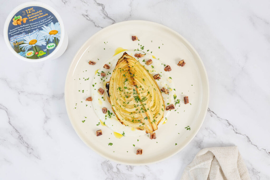 Kabisspalten auf Mandel-Joghurt-Sauce mit Pekannüssen und Petersilie, daneben Becher veganer Mandeljoghurt