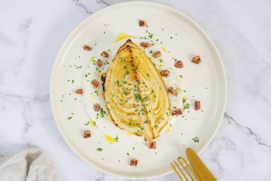 Nahaufnahme Ofenkohlspalten auf Joghurtsauce mit Petersilie und gebrannten Nüssen, Besteck am Teller