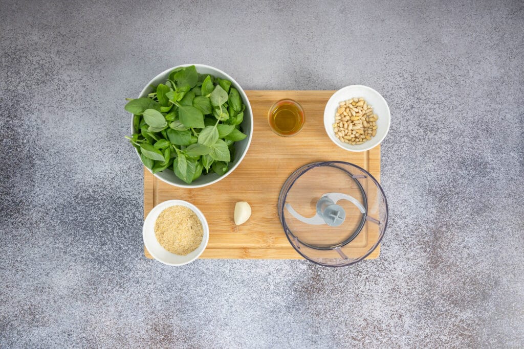 Vorbereitung für Basilikum-Pesto ohne Parmesan: gepflückter Basilikum, Pinienkerne, Knoblauch und Olivenöl beim Food Processor auf Holzbrett