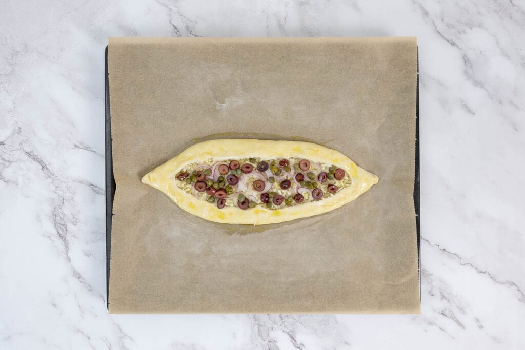 Ungebackenes Pide mit Oliven, Kapern und Zwiebeln auf dem Backblech – vor dem Backen mit Olivenöl bestrichen