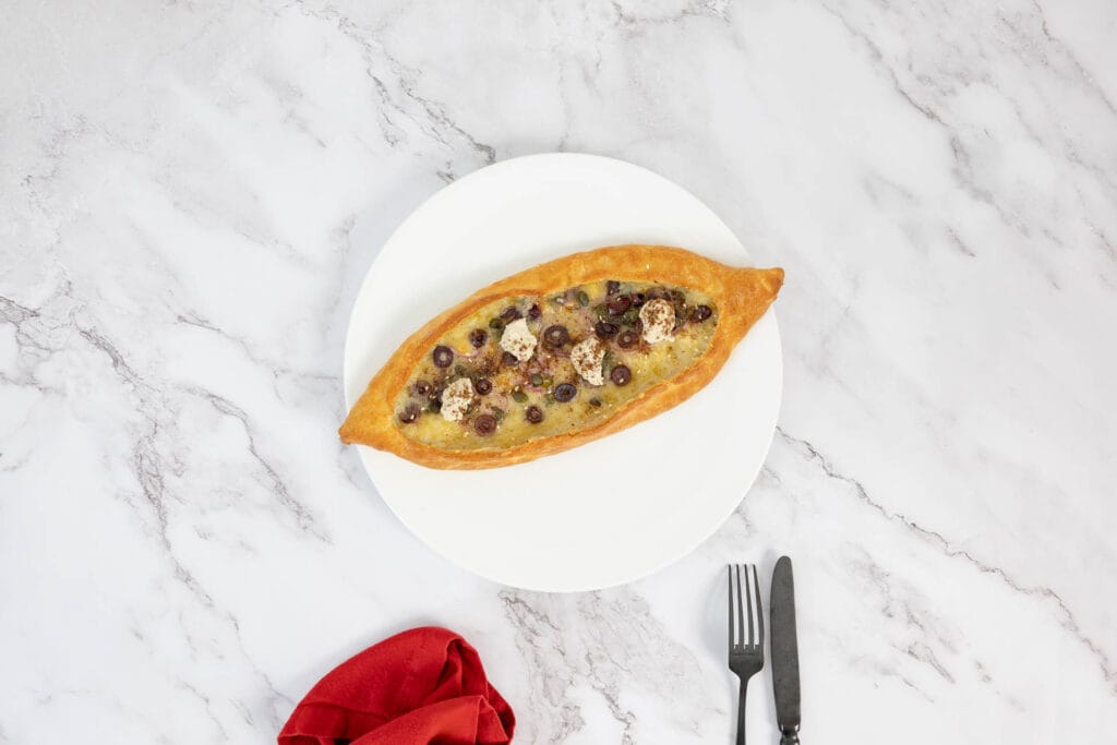 Pide mit Oliven, Zaatar und Ricotta auf Teller mit roter Serviette und schwarzem Besteck – moderne Foodfotografie