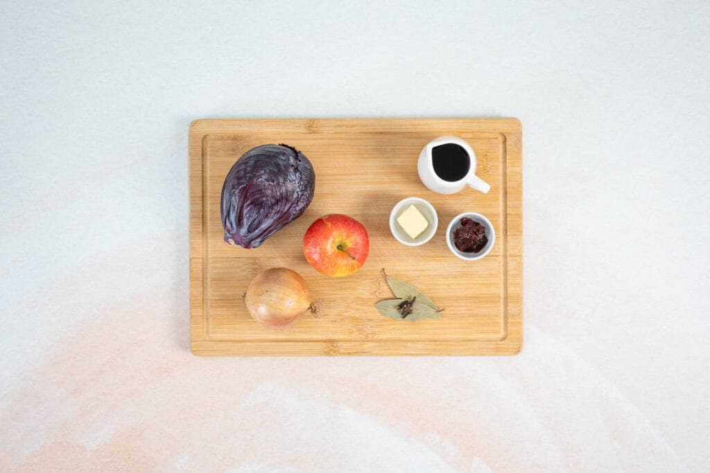 Zutaten für klassisches veganes Rotkraut mit Rotkohl, Apfel, Zwiebel, Rotwein, Gewürzen und Preiselbeerkonfitüre auf einem Holzbrett