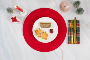 Hand hebt den Teller mit veganer Morchelterrine, Stern-Toast und Preiselbeeren von der festlichen Weihnachtstafel an.