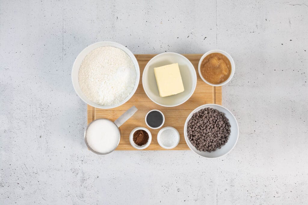 Zutaten für vegane Chocolate Chip Cookies – Mehl, vegane Butter, Apfelmus, Zucker, Melasse, Schokodrops und Meersalz auf einem Holzbrett