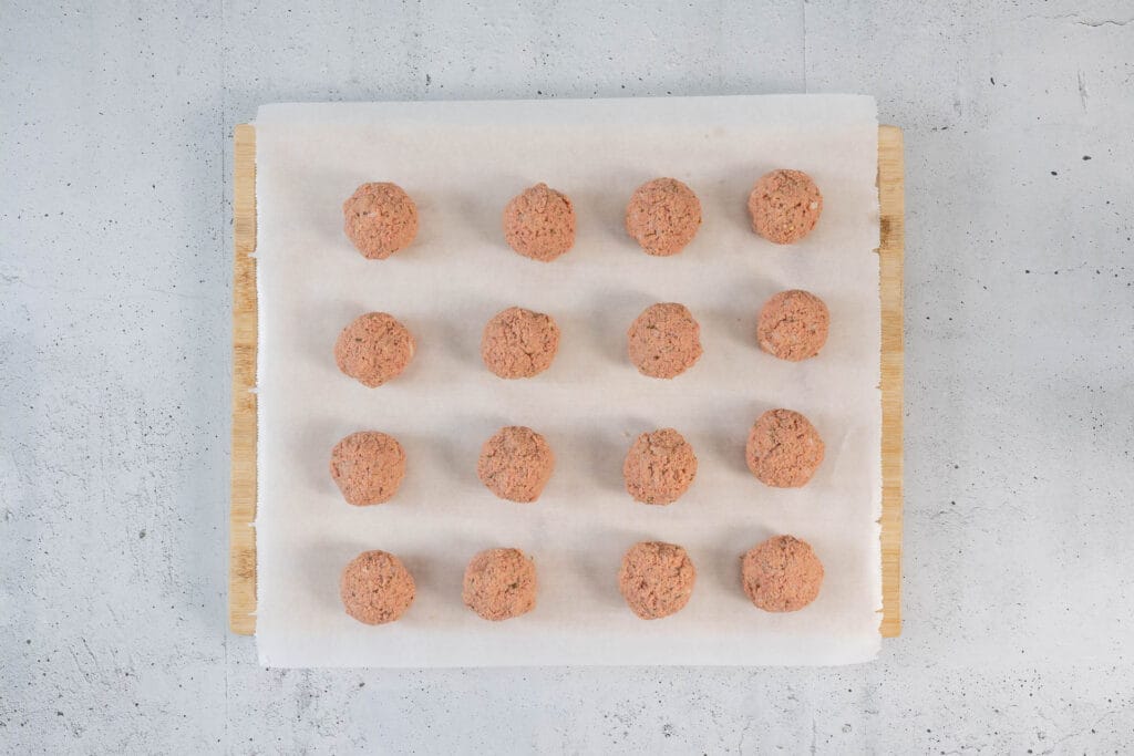Sechzehn geformte vegane Klopse auf einem mit Backpapier ausgelegten Holzbrett, bereit zum Garen