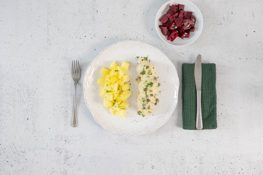 Vegane Königsberger Klopse mit Kapernsauce und Salzkartoffeln auf dekoriertem Teller, dazu Randensalat und grüne Serviette