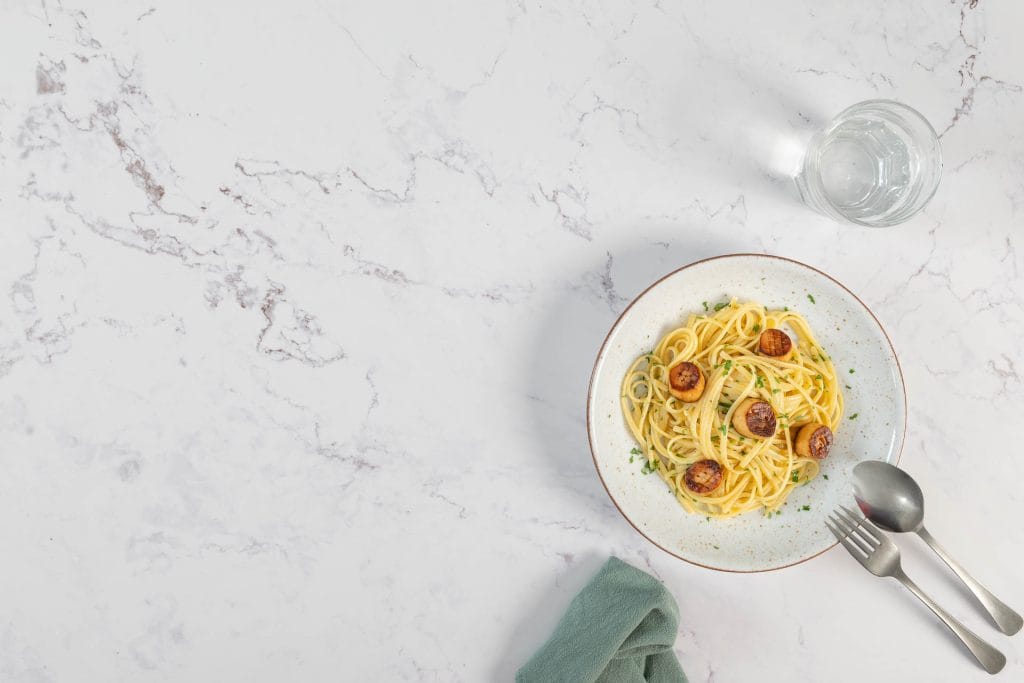 Vegane Muschelpasta mit goldbraunen Pilz-Jakobsmuscheln auf einem hellen Keramikteller, daneben Wasserglas und Besteck