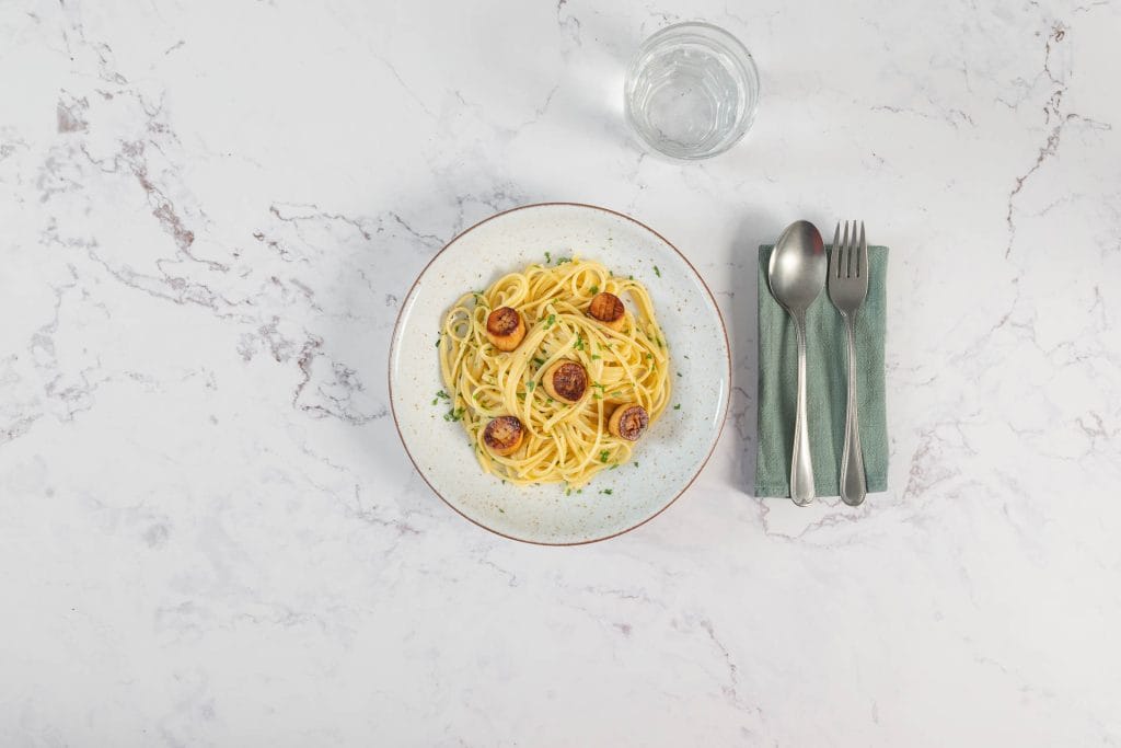Linguine mit veganen Jakobsmuscheln aus Kräuterseitlingen, serviert mit Stoffserviette und Besteck auf weissem Marmor