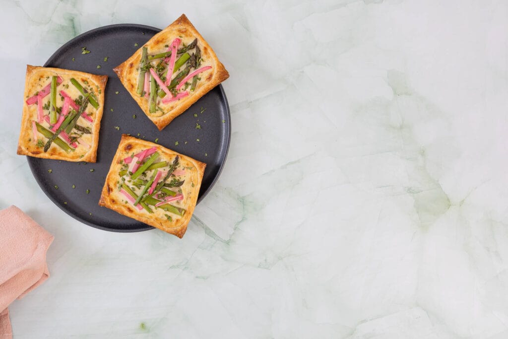 Drei Spargel-Rhabarber-Tartelettes mit Blätterteig und veganem Frischkäse auf dunklem Teller serviert