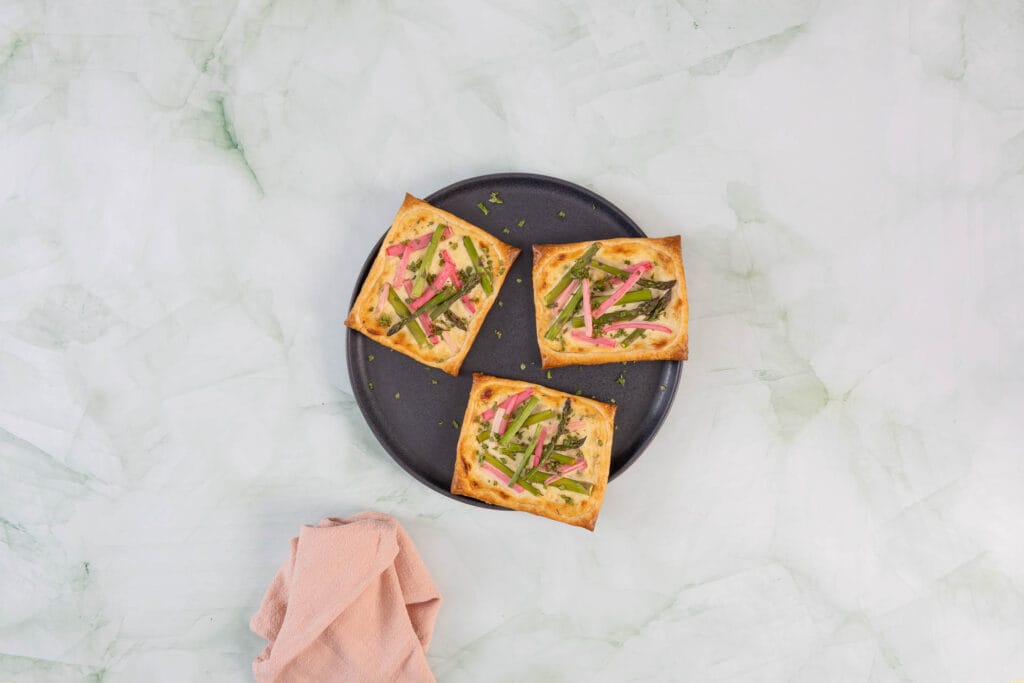 Drei vegane Tartelettes mit Spargel und Rhabarber auf dunklem Teller, garniert mit frischer Minze – Frühlingsrezept