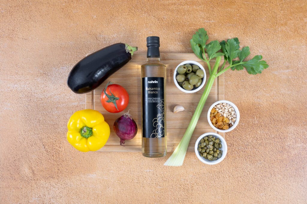 Zutaten für vegane Caponata auf einem Holzbrett: Aubergine, Tomate, gelbe Peperoni, rote Zwiebel, Knoblauch, Stangensellerie, grüne Oliven, Kapern, Rosinen, Pinienkerne und Balsamico Bianco