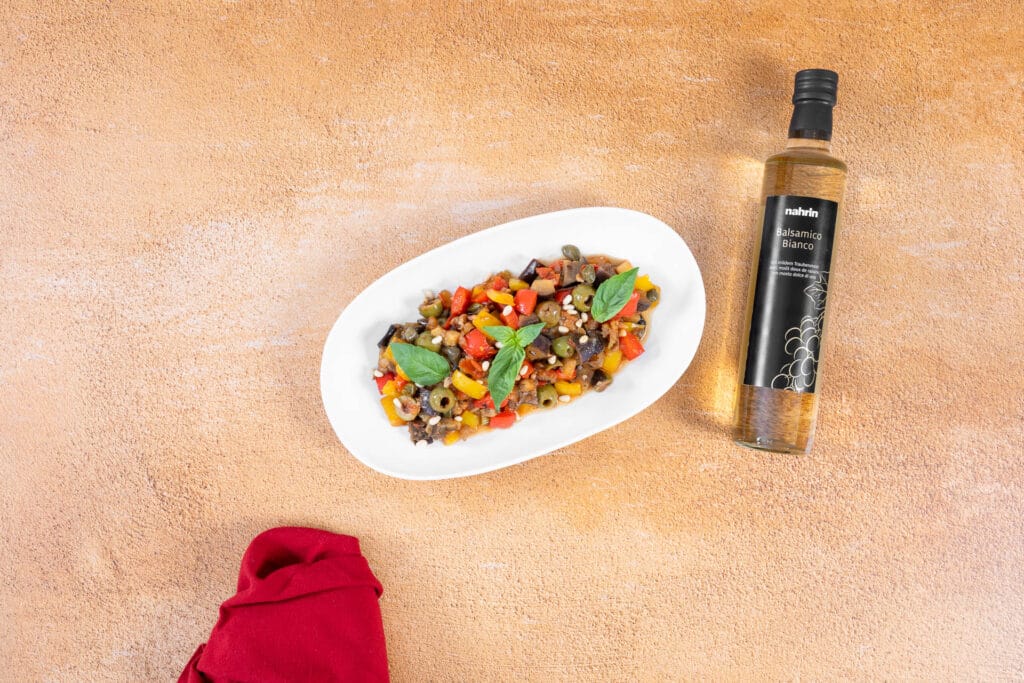 Angerichtete Caponata mit Agrodolce auf einer weissen Platte, garniert mit Basilikum, daneben eine Flasche Balsamico Bianco