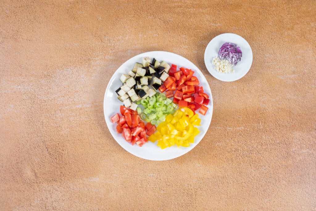 Klein gewürfeltes Gemüse für die sizilianische Caponata auf einem Teller: Aubergine, rote und gelbe Peperoni, Tomaten und Sellerie, daneben gehackte Zwiebel und Knoblauch