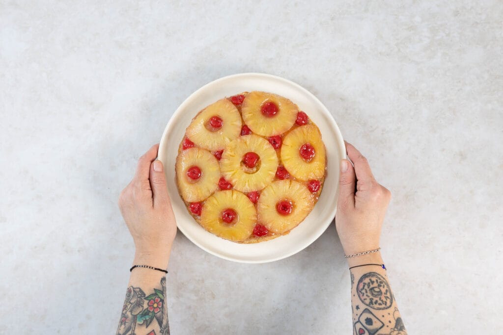 Zwei Hände halten den fertigen veganen Ananas Upside Down Cake auf einer Tortenplatte