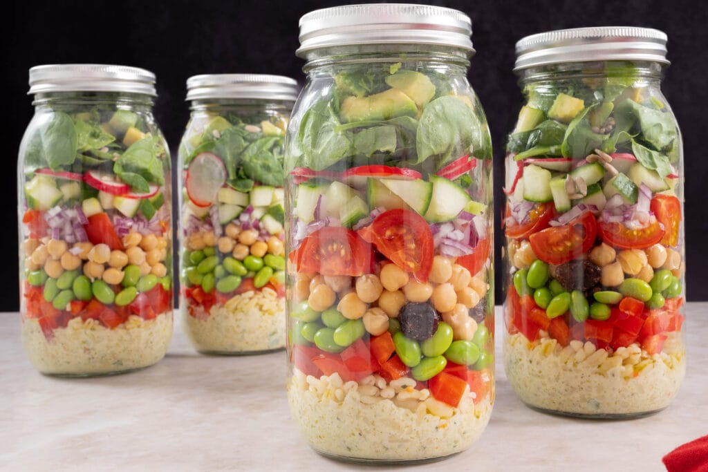 Vier vegane Meal-Prep-Salate im Glas mit Deckel, bereit für das Büro