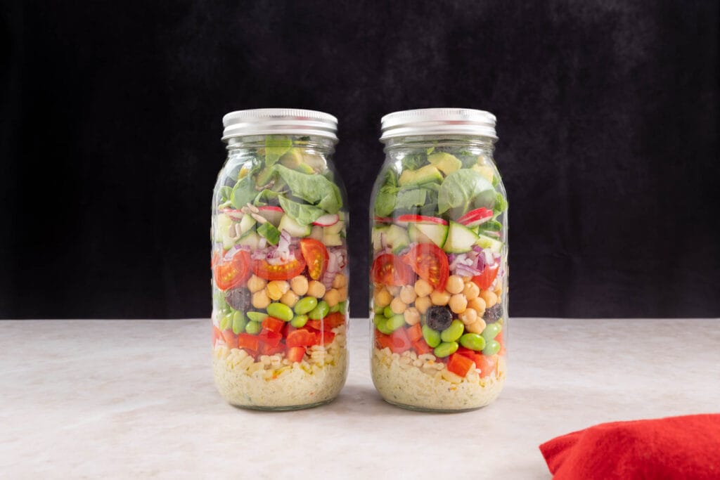 Zwei fertige vegane Salate im Glas zum Mitnehmen mit bunten Gemüseschichten