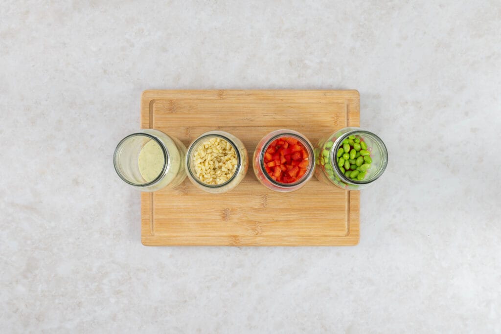 Veganer Salat im Glas beim Schichten – Dressing, Ebly, Peperoni und Edamame als unterste Schichten
