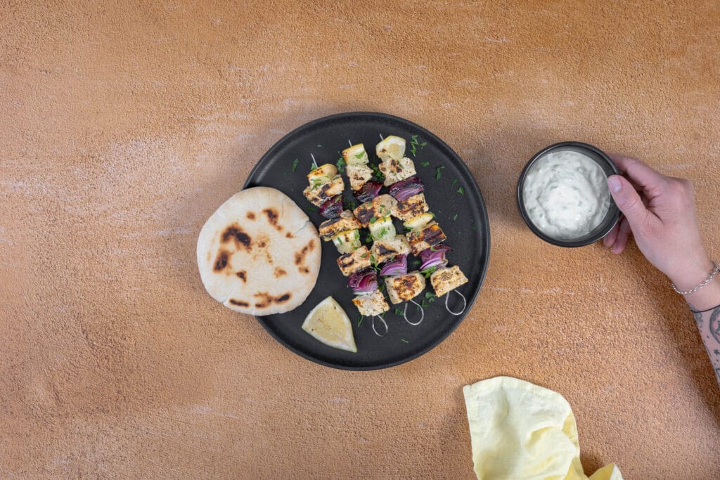 Hand greift zur Schale mit veganem Tzatziki neben einem Teller mit Souvlaki und Pitabrot
