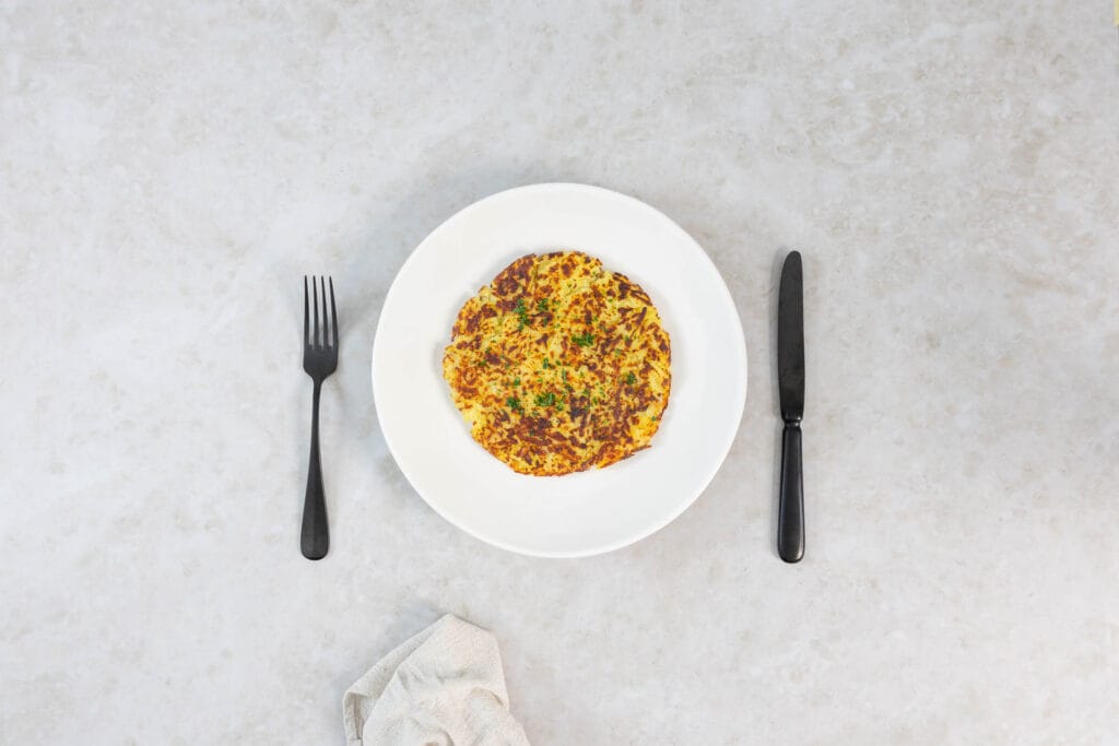 Knusprige vegane Rösti auf weissem Teller mit dunklem Besteck angerichtet