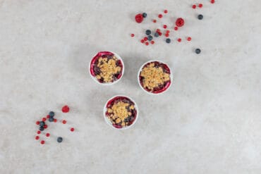 Drei Portionen Beeren-Crumble mit knuspriger Haferflocken-Kruste, umgeben von frischen Sommerbeeren