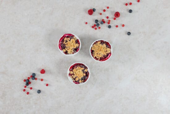 Drei Portionen Beeren-Crumble mit knuspriger Haferflocken-Kruste, umgeben von frischen Sommerbeeren
