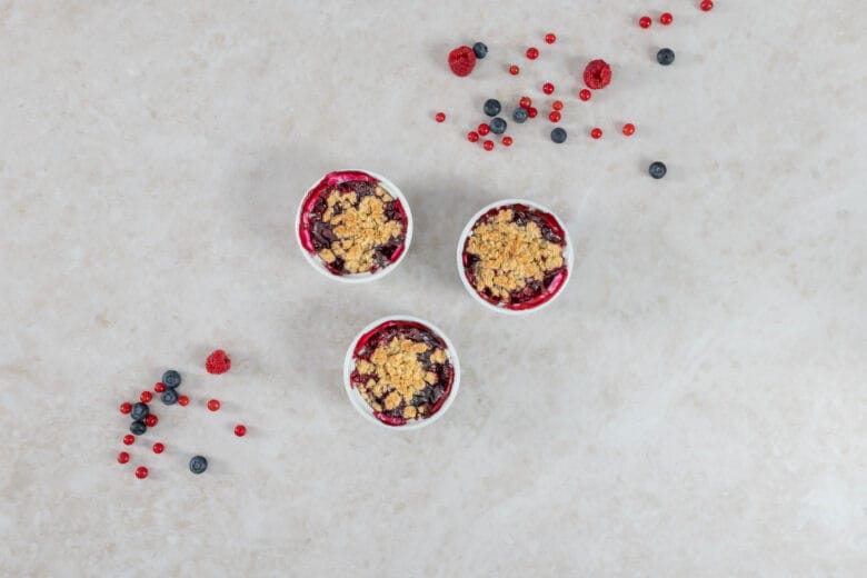 Drei Portionen Beeren-Crumble mit knuspriger Haferflocken-Kruste, umgeben von frischen Sommerbeeren