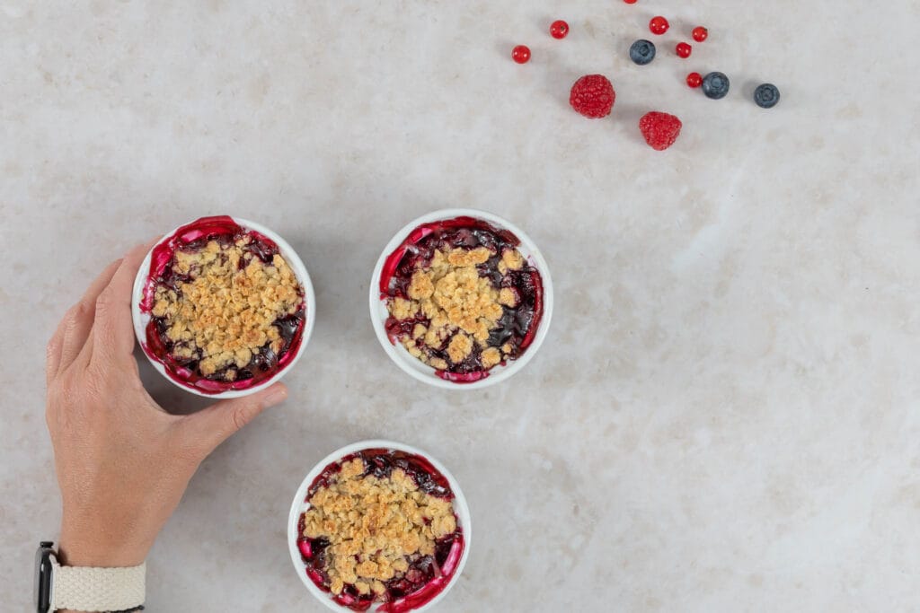 Hand hält ein Ofenschälchen mit warmem veganem Beeren-Crumble, zwei weitere Portionen daneben