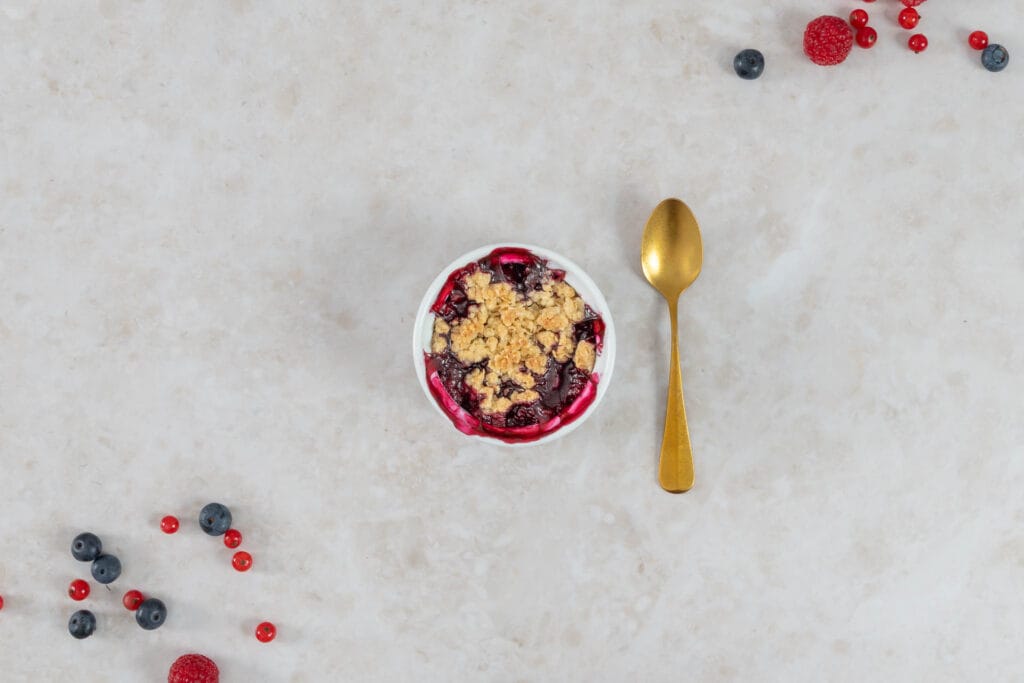 Veganer Beeren-Crumble mit Haferflocken-Streuseln, goldenem Löffel und frischen Beeren angerichtet