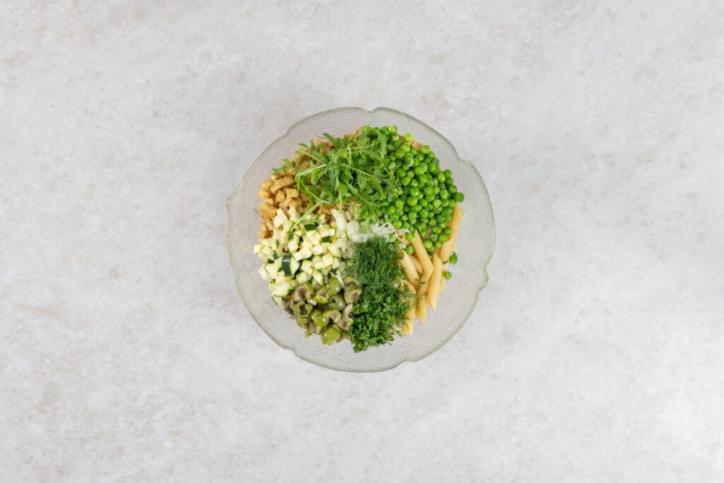 Penne mit Erbsen, Zucchetti, Oliven, Rucola und Kräutern in einer Schüssel vor dem Vermengen