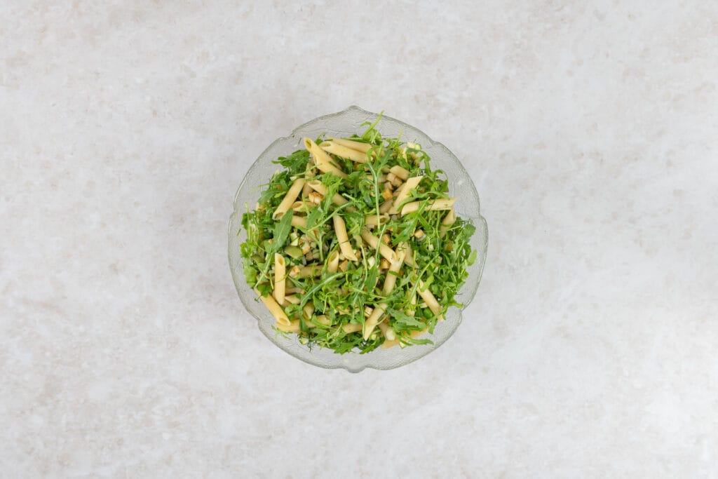 Grüner Pastasalat mit Rucola und Penne in einer Glasschüssel, mit dem Dressing vermengt