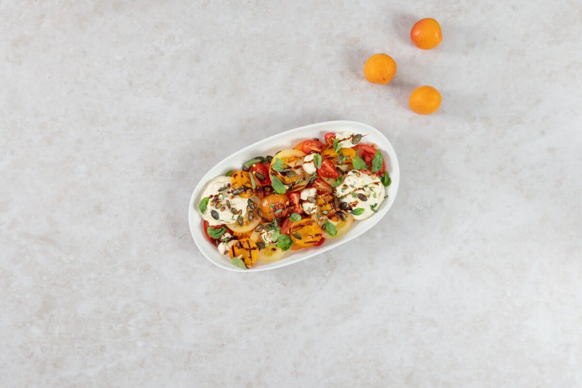 Grillierter Aprikosensalat mit gegrillten Aprikosen, bunten Tomaten und veganer Ricottacreme auf einer weissen Platte, daneben frische Aprikosen