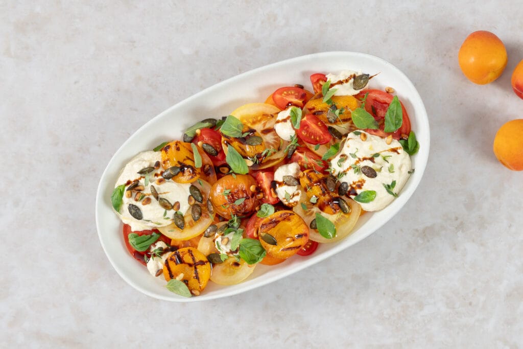 Grillierter Aprikosensalat mit gegrillten Aprikosen, bunten Tomaten und veganer Ricottacreme, angerichtet mit frischen Kräutern und frischen Aprikosen
