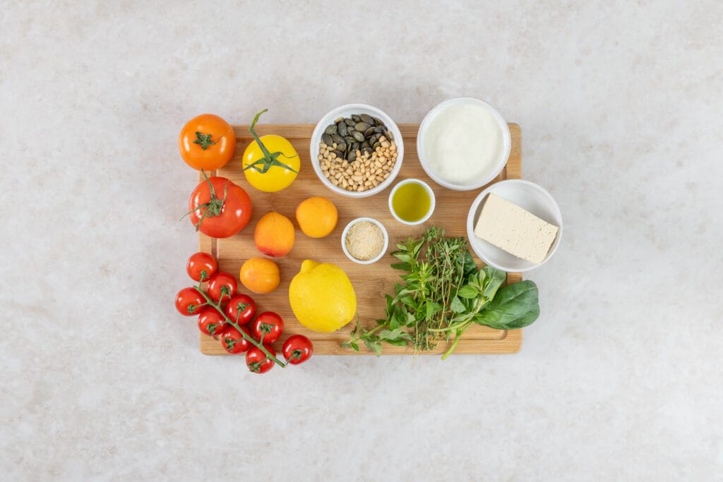 Zutaten für grillierten Aprikosensalat auf einem Holzbrett: bunte Tomaten, Aprikosen, Tofu, veganer Joghurt, Zitrone, Kürbis- und Pinienkerne sowie frische Kräuter