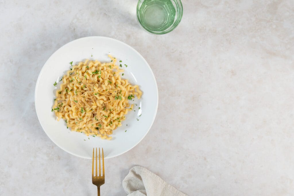 Cremige Zwiebel-Pasta mit Petersilie, daneben ein Glas Wasser auf hellem Steinuntergrund