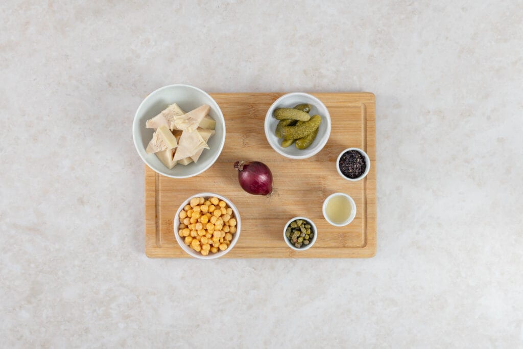 Veganer Thonsalat, die Zutaten: Jackfruit, Kichererbsen, rote Zwiebel, Cornichons, Kapern, Kapernwasser und Noriflocken auf einem Holzbrett