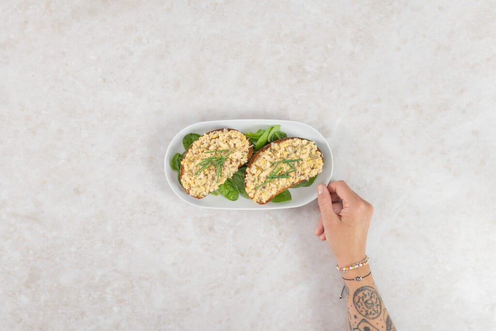 Hand hält eine Platte, darauf veganer Thonsalat auf Ruchbrot mit frischem Spinat
