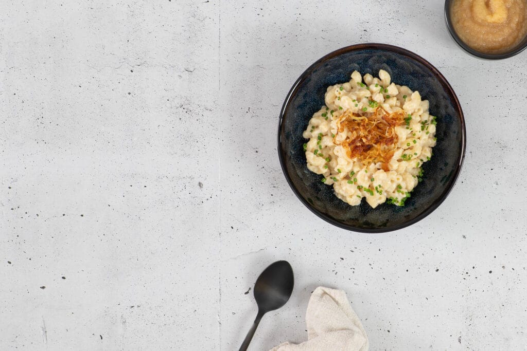 Cremige Hörnli mit veganer Käsesauce und goldbraunen Röstzwiebeln, im Hintergrund Apfelmus