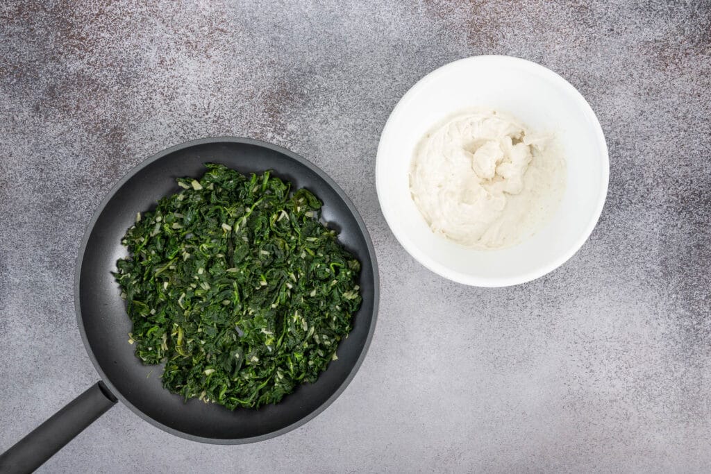 Gedünsteter Spinat mit Zwiebeln in der Bratpfanne und veganer Frischkäse mit Hefeflocken in einer Schüssel